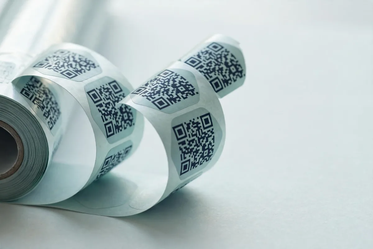 A roll of die-cut stickers peeling from its backing paper across a pale desk, every sticker printed with the same QR code.