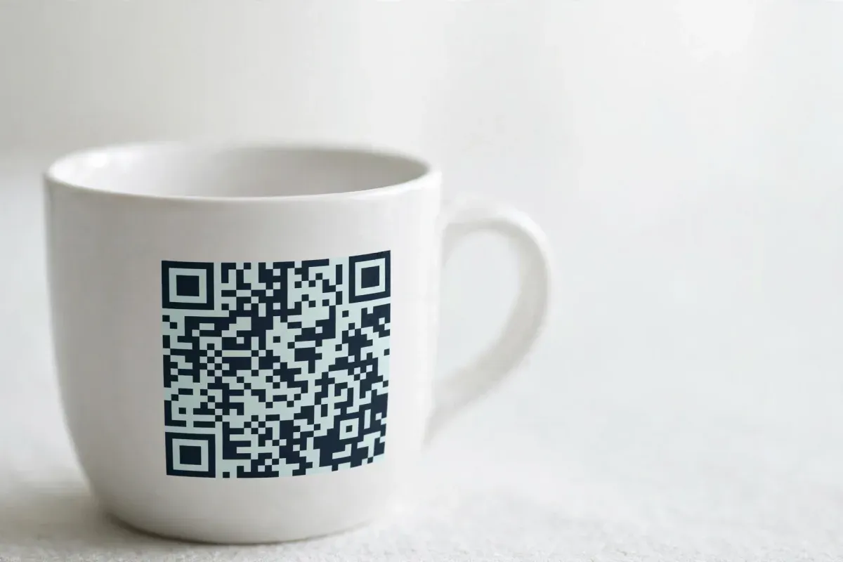 A plain white ceramic mug printed with a small QR code, on a pale table.