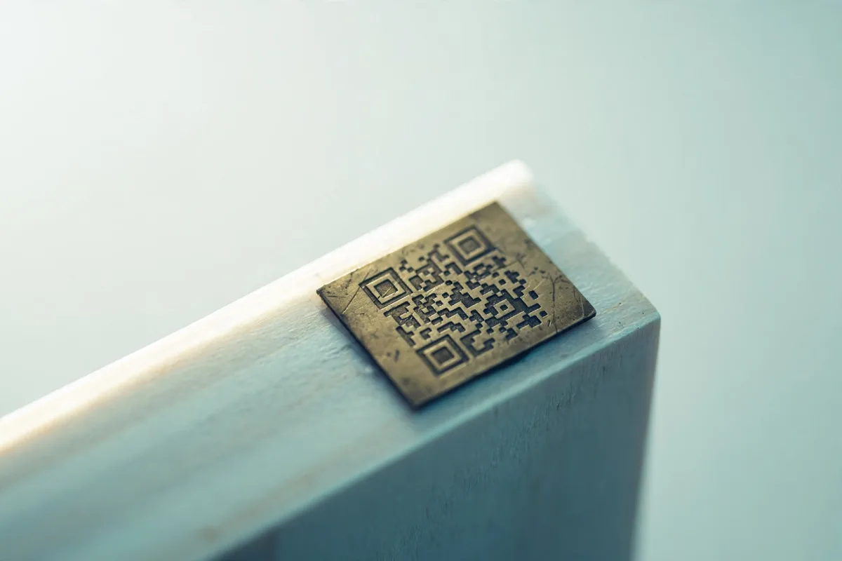 A small brass plaque etched with a QR code mounted on a pale wooden plinth, seen at a slight angle.
