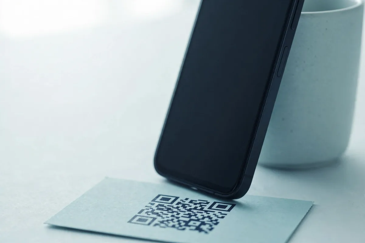 A smartphone propped upright against a pale ceramic mug on a desk, its screen dark, a postcard printed with a QR code lying flat in front of it.