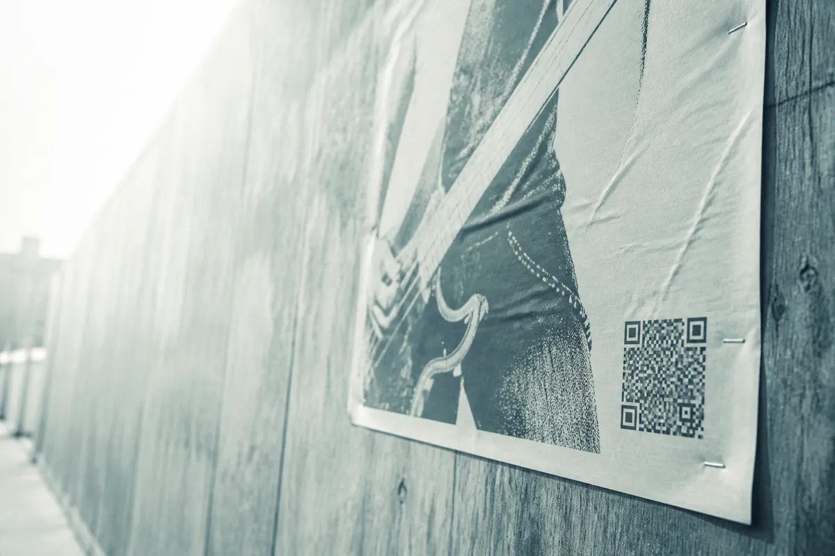 A paper poster pasted flat to a plywood hoarding and photographed along the wall, a QR code printed low on the sheet.