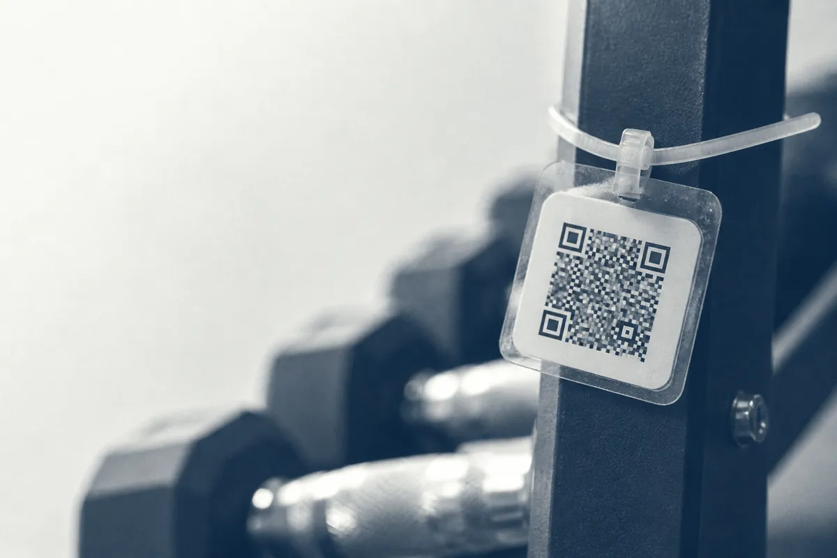 A laminated tag zip-tied to the upright of a dumbbell rack, a QR code printed on it, weights out of focus behind.
