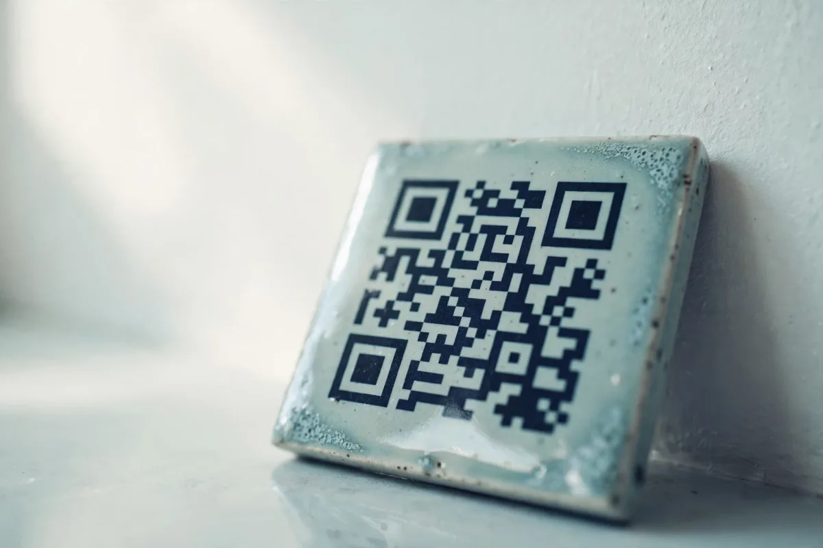 A glazed ceramic tile printed with a QR code leaning against a pale plaster wall, its reflection faint on the glaze.