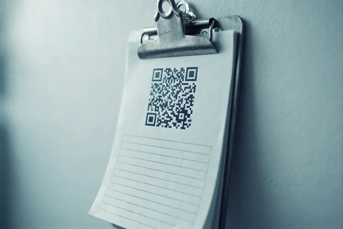 A metal clipboard on a pale workshop wall, its sheet printed with a QR code above a blank ruled area.