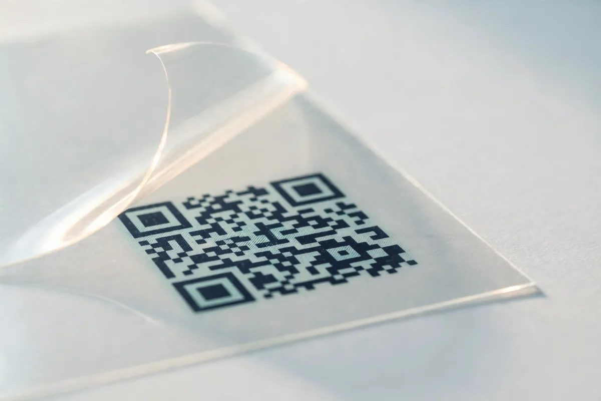 A sheet of clear acetate printed with a QR code lying over a pale desk, one corner lifted so light catches the sheet.