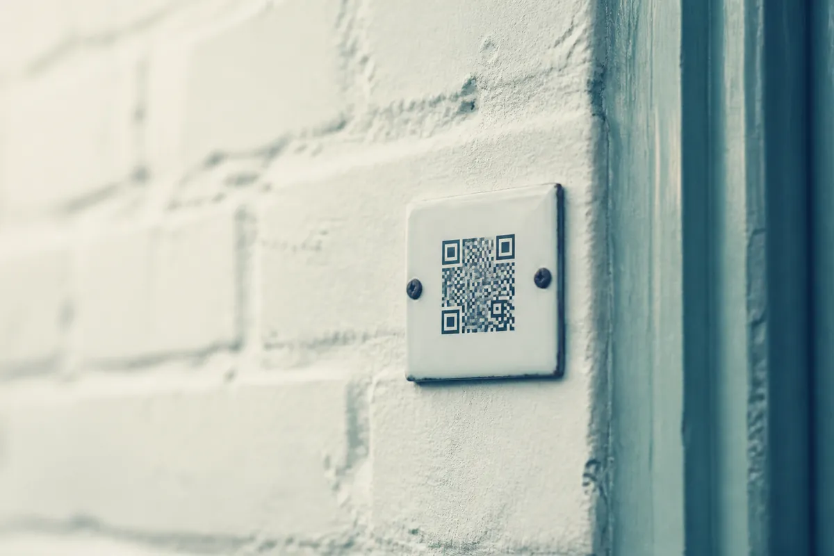 A pale enamel plate printed with a QR code mounted on a painted brick wall beside a doorframe.