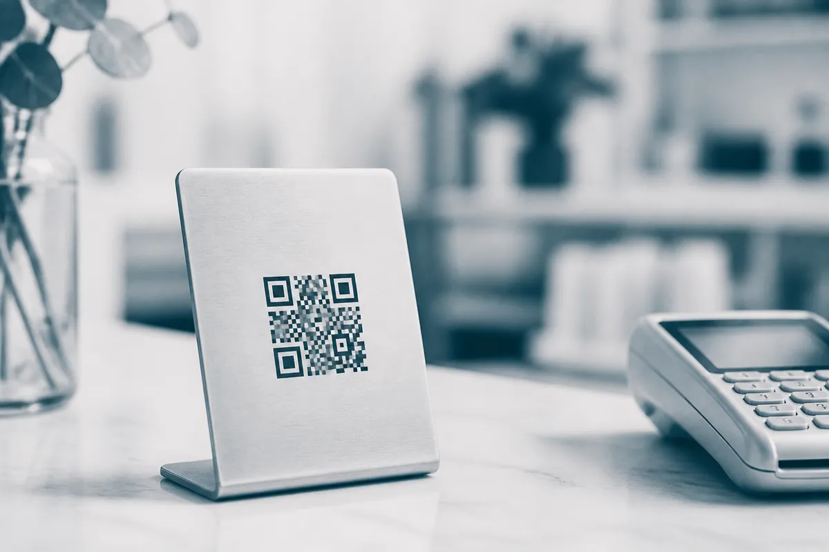 A small sign printed with a QR code in a metal holder on a shop counter beside a card reader.